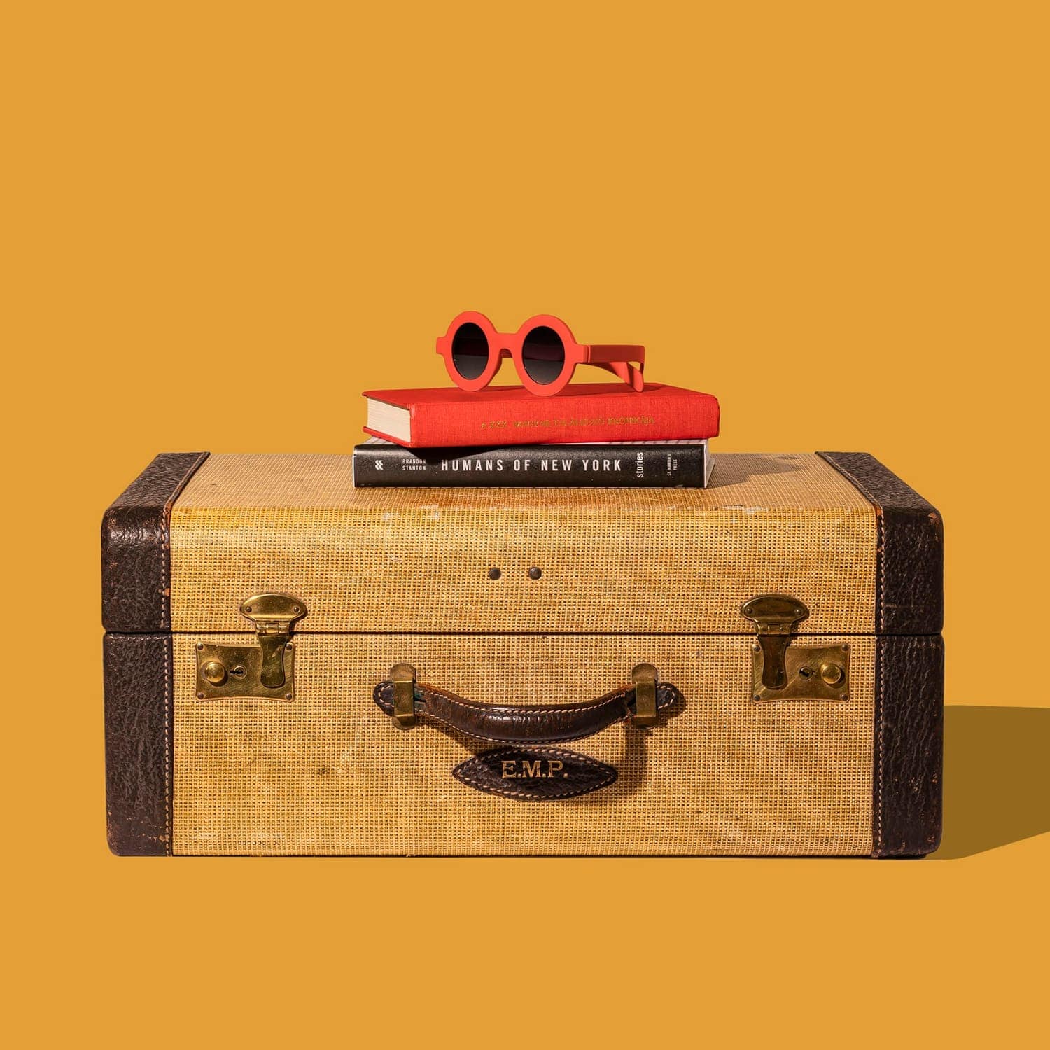 A vintage suitcase topped with books and red sunglasses on an orange background
