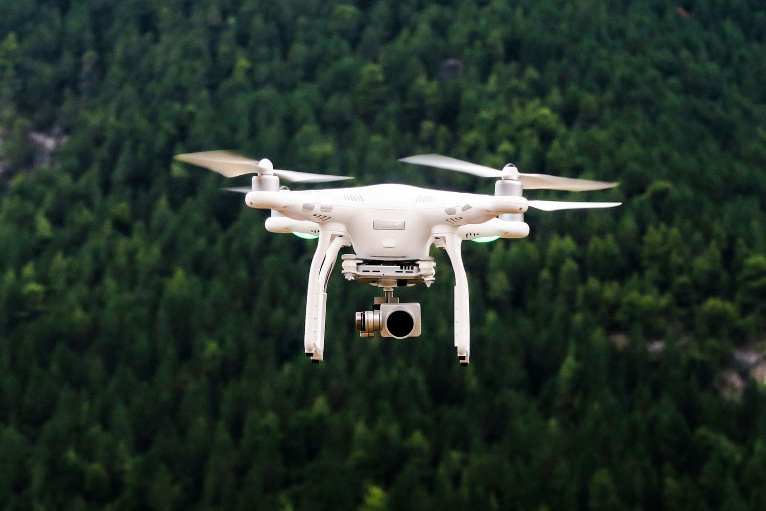 White camera drone flying over a green forest